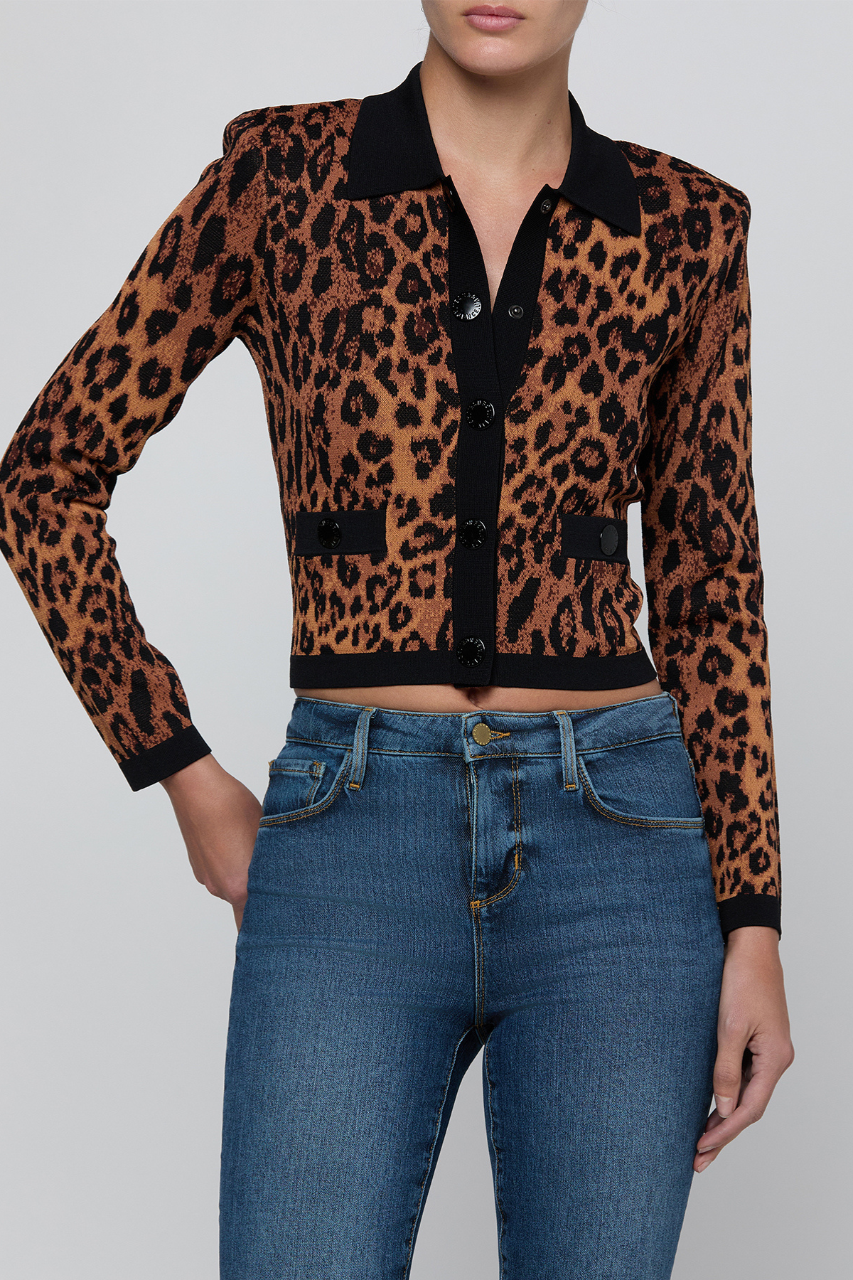 A woman wearing a cropped leopard-print cardigan with a black collar and front buttons, paired with blue jeans, against a light gray studio background.