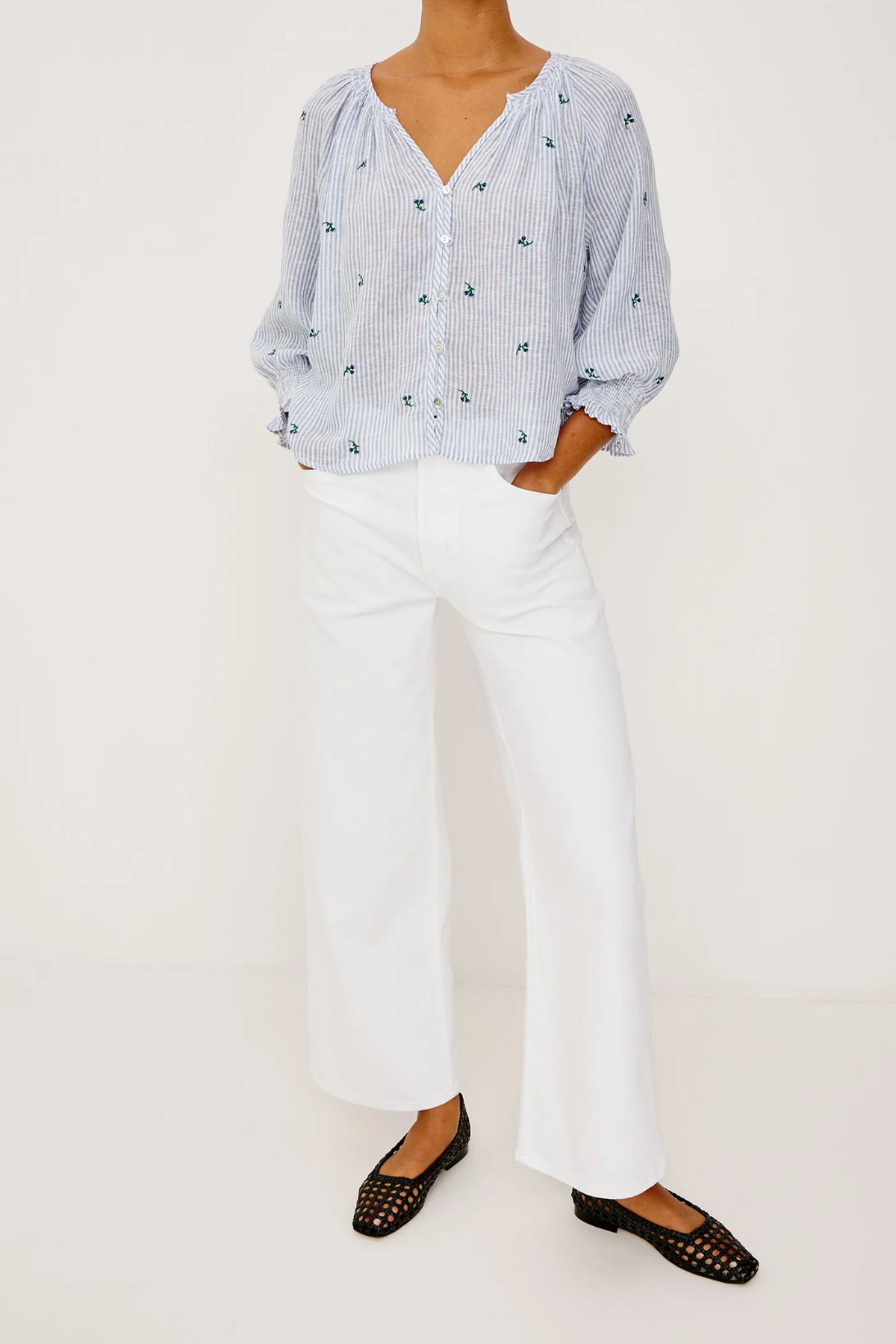 Person wearing a blue patterned blouse, white pants, and black flats on a white background