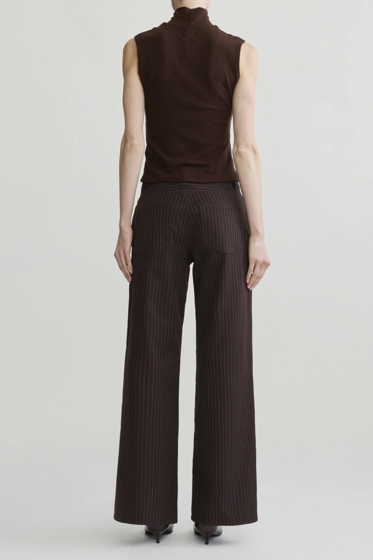 A model shown from the back wearing a sleeveless dark brown mock-neck top and dark brown pinstriped wide-leg trousers, set against a plain light gray studio background.