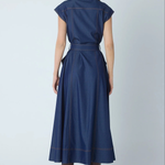 A woman viewed from the back wearing a navy blue midi dress with cap sleeves, a matching waist tie, and contrasting brown contrast stitching, paired with brown heeled boots against a light gray studio background.