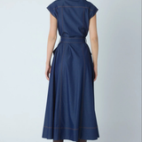 A woman viewed from the back wearing a navy blue midi dress with cap sleeves, a matching waist tie, and contrasting brown contrast stitching, paired with brown heeled boots against a light gray studio background.