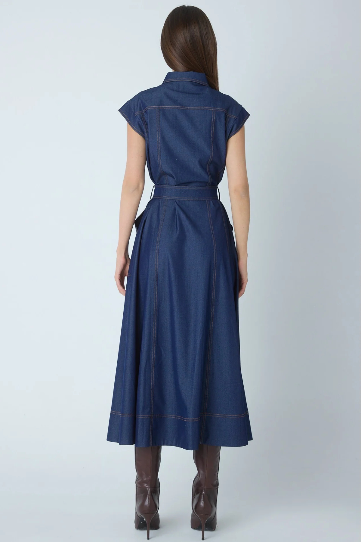 A woman viewed from the back wearing a navy blue midi dress with cap sleeves, a matching waist tie, and contrasting brown contrast stitching, paired with brown heeled boots against a light gray studio background.