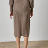 A model from the back wearing a taupe ribbed knit midi skirt and matching knit sweater, paired with tan heeled sandals, set against a neutral studio backdrop.
