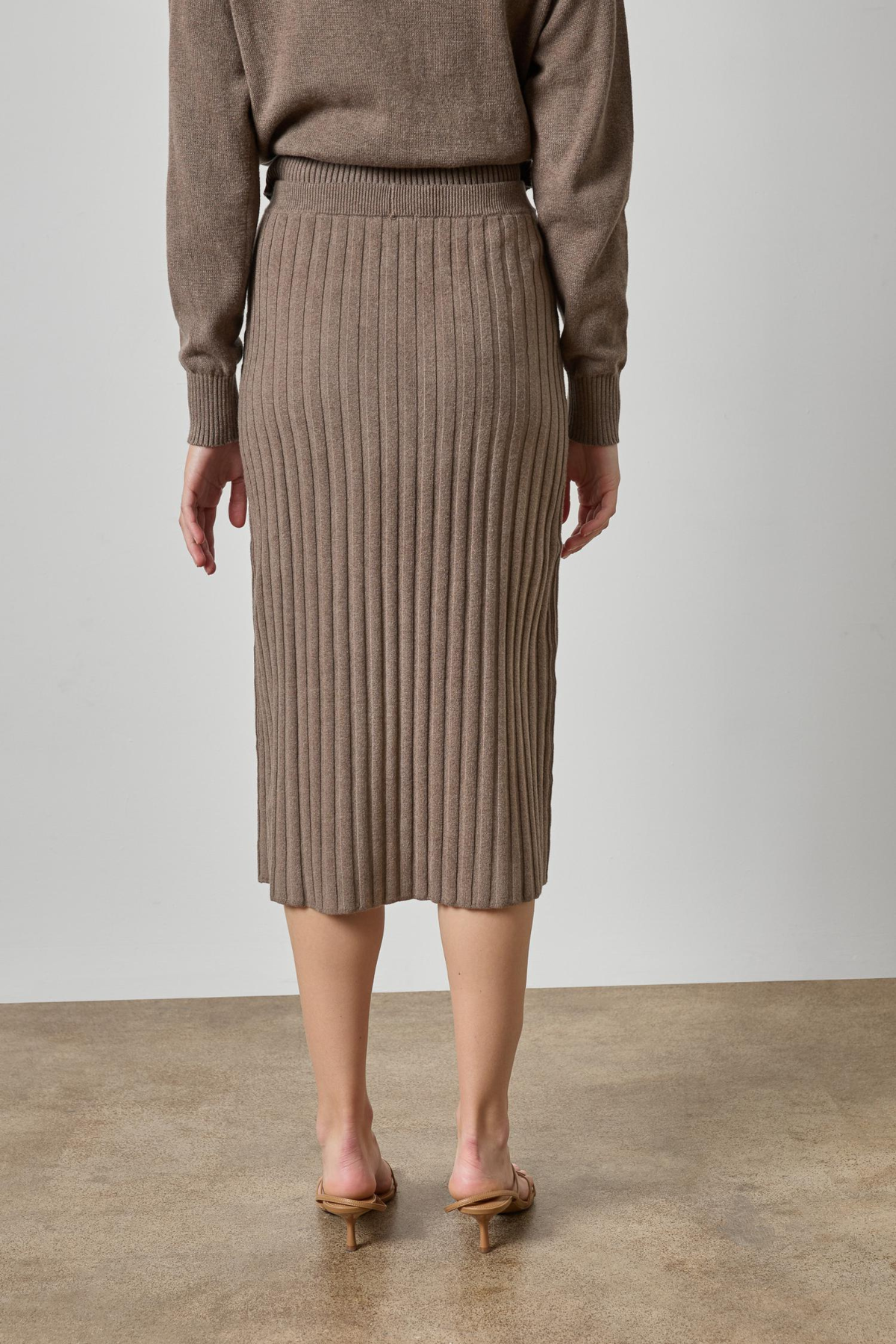 A model from the back wearing a taupe ribbed knit midi skirt and matching knit sweater, paired with tan heeled sandals, set against a neutral studio backdrop.