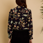 A woman viewed from the back wearing a long-sleeve button-up blouse with a dark navy floral print and black pants, set against a warm beige background with a plant leaf visible on the right.