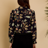 A woman viewed from the back wearing a long-sleeve button-up blouse with a dark navy floral print and black pants, set against a warm beige background with a plant leaf visible on the right.