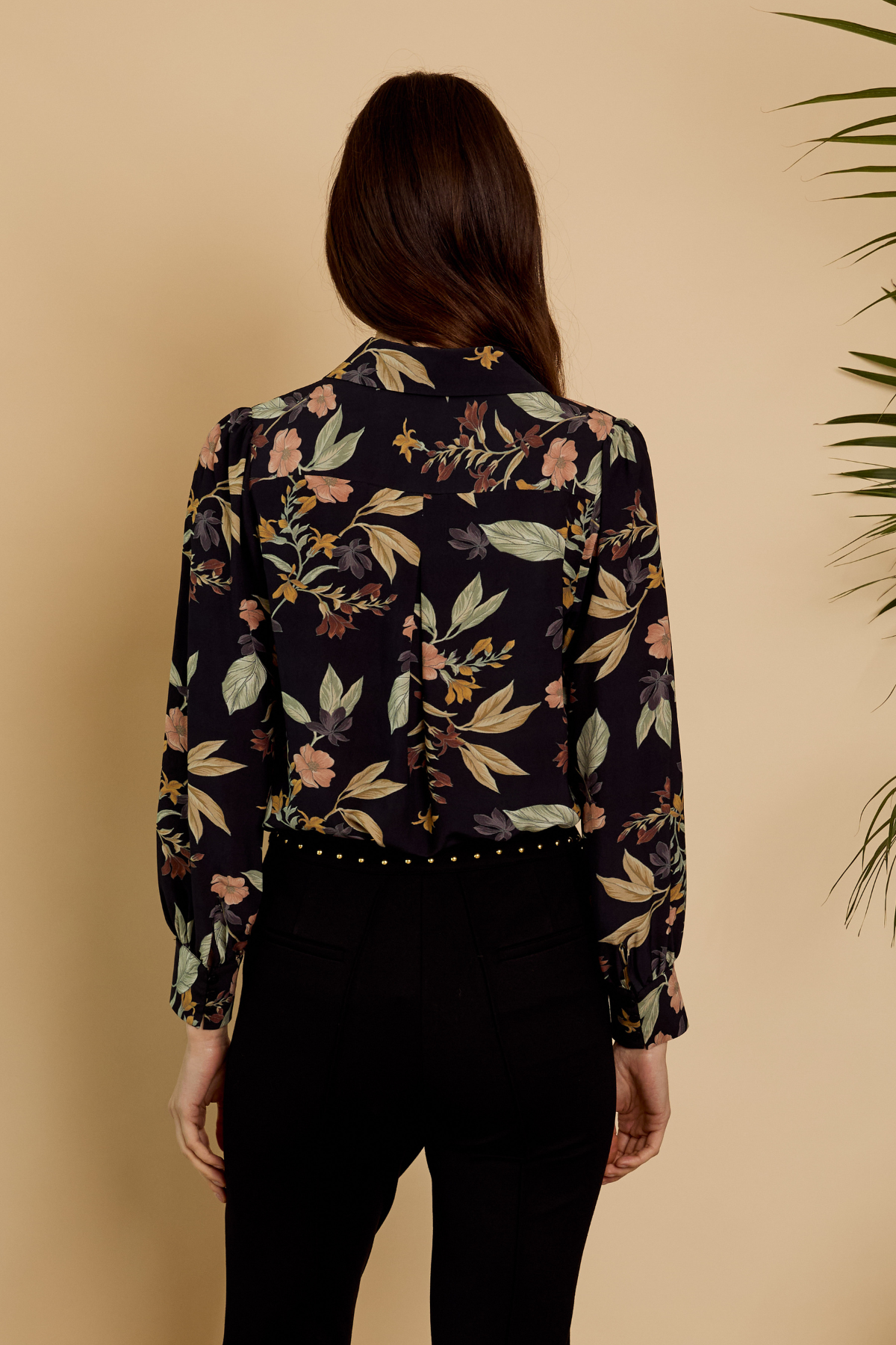 A woman viewed from the back wearing a long-sleeve button-up blouse with a dark navy floral print and black pants, set against a warm beige background with a plant leaf visible on the right.