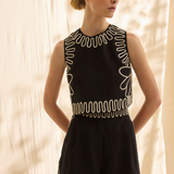 A woman modeling a sleeveless black romper with white embroidered geometric and floral patterns, set against a warm beige backdrop with soft leaf shadows.