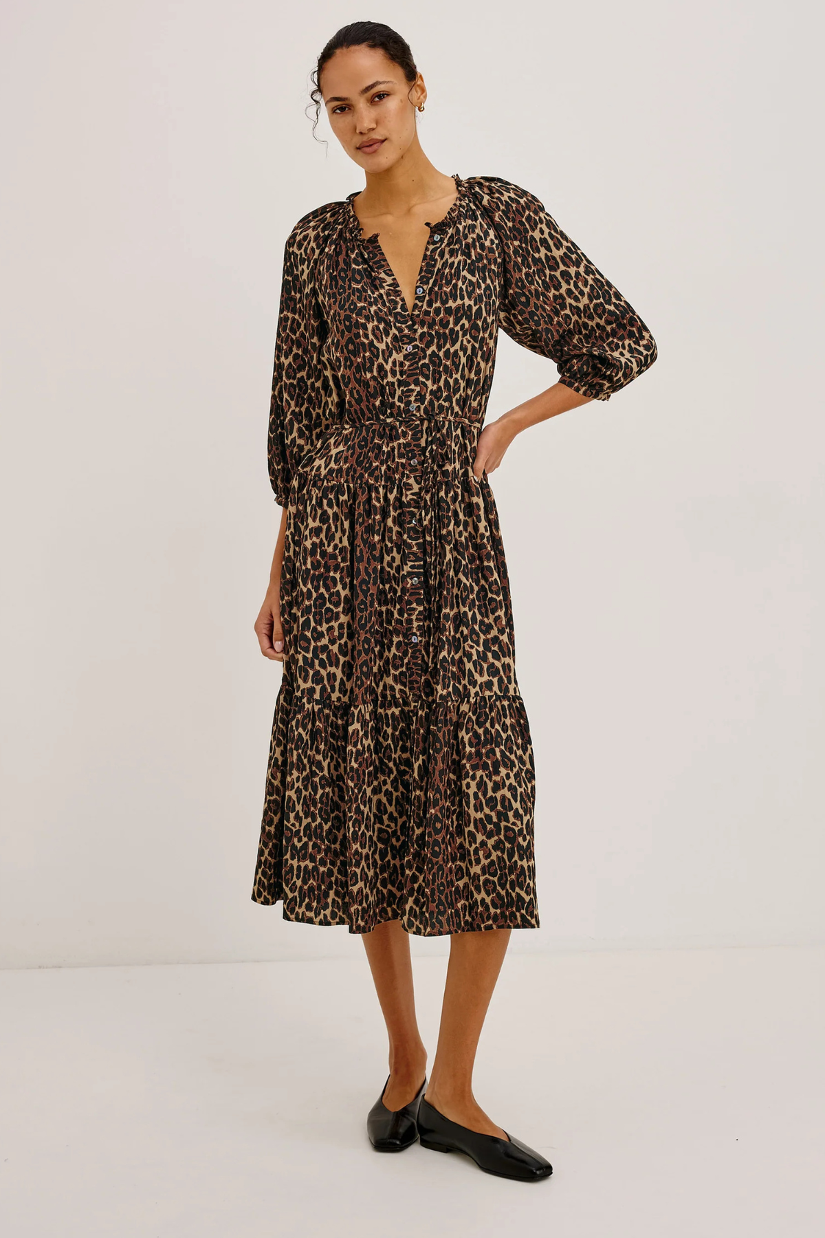 A woman modeling a leopard-print midi dress with three-quarter sleeves and a V-neckline, paired with black flats, against a plain white studio background.