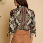 A woman shown from the back wearing a long-sleeve button-up shirt with a detailed botanical and bird pattern in brown, green, and beige, paired with brown trousers, set against a warm beige background.