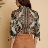 A woman shown from the back wearing a long-sleeve button-up shirt with a detailed botanical and bird pattern in brown, green, and beige, paired with brown trousers, set against a warm beige background.