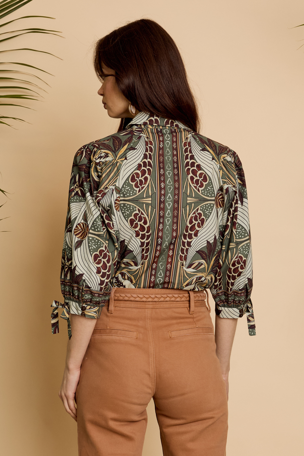 A woman shown from the back wearing a long-sleeve button-up shirt with a detailed botanical and bird pattern in brown, green, and beige, paired with brown trousers, set against a warm beige background.