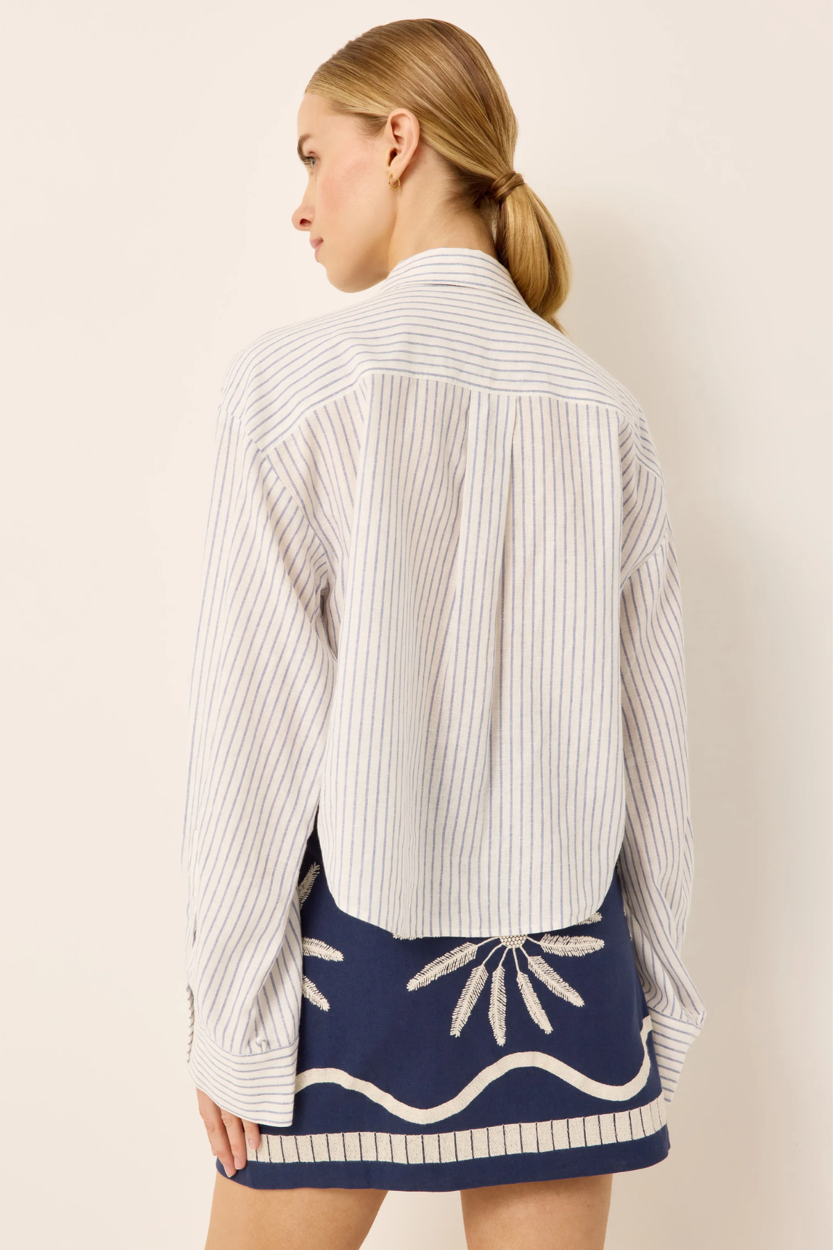 A woman shown from the back wearing a white long-sleeve button-up shirt with thin vertical navy stripes and a navy mini skirt with white embroidered details, against a plain beige background.