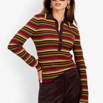 A woman modeling a long-sleeve knit polo-style top with horizontal stripes in red, yellow, green, and brown over a dark base, paired with dark brown pants against a light gray studio background.