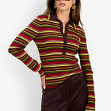 A woman modeling a long-sleeve knit polo-style top with horizontal stripes in red, yellow, green, and brown over a dark base, paired with dark brown pants against a light gray studio background.