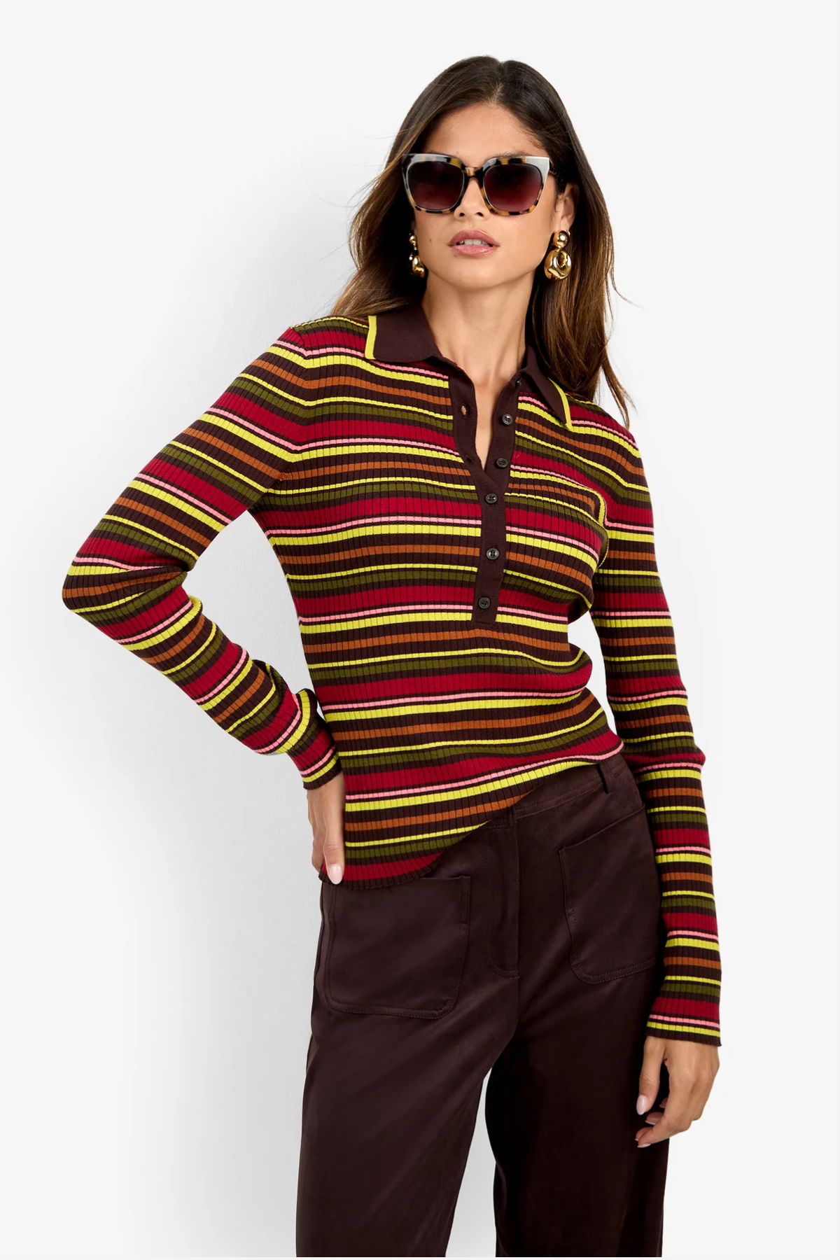 A woman modeling a long-sleeve knit polo-style top with horizontal stripes in red, yellow, green, and brown over a dark base, paired with dark brown pants against a light gray studio background.