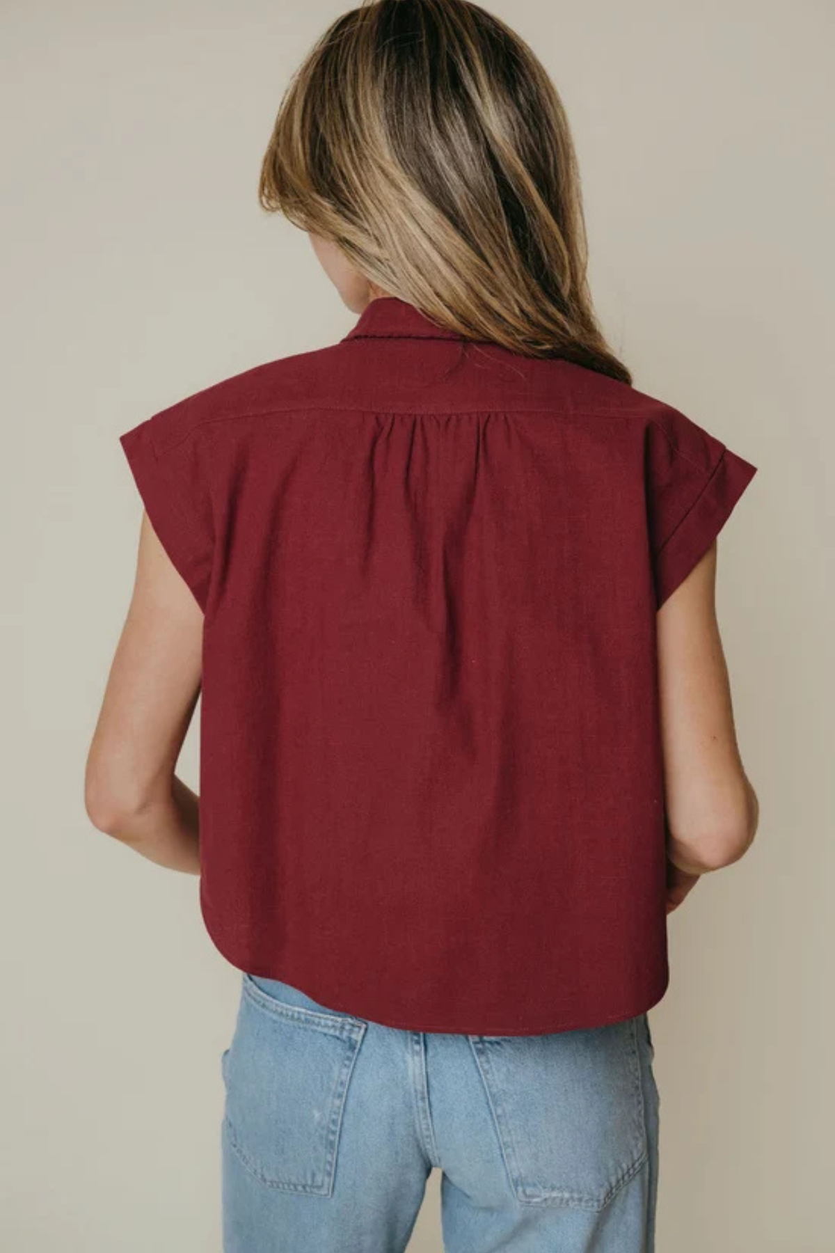 A woman shown from behind wearing a sleeveless, deep burgundy top and light blue jeans against a plain beige background.