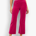 A model wearing bright fuchsia pink velvet cropped flare pants, paired with a white ribbed top and white heeled sandals, standing against a white studio background.