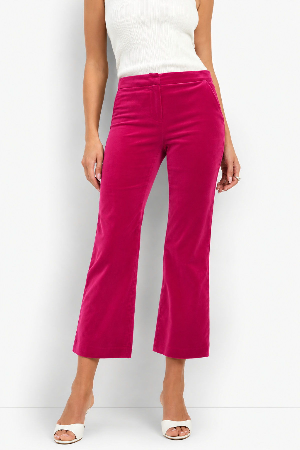 A model wearing bright fuchsia pink velvet cropped flare pants, paired with a white ribbed top and white heeled sandals, standing against a white studio background.