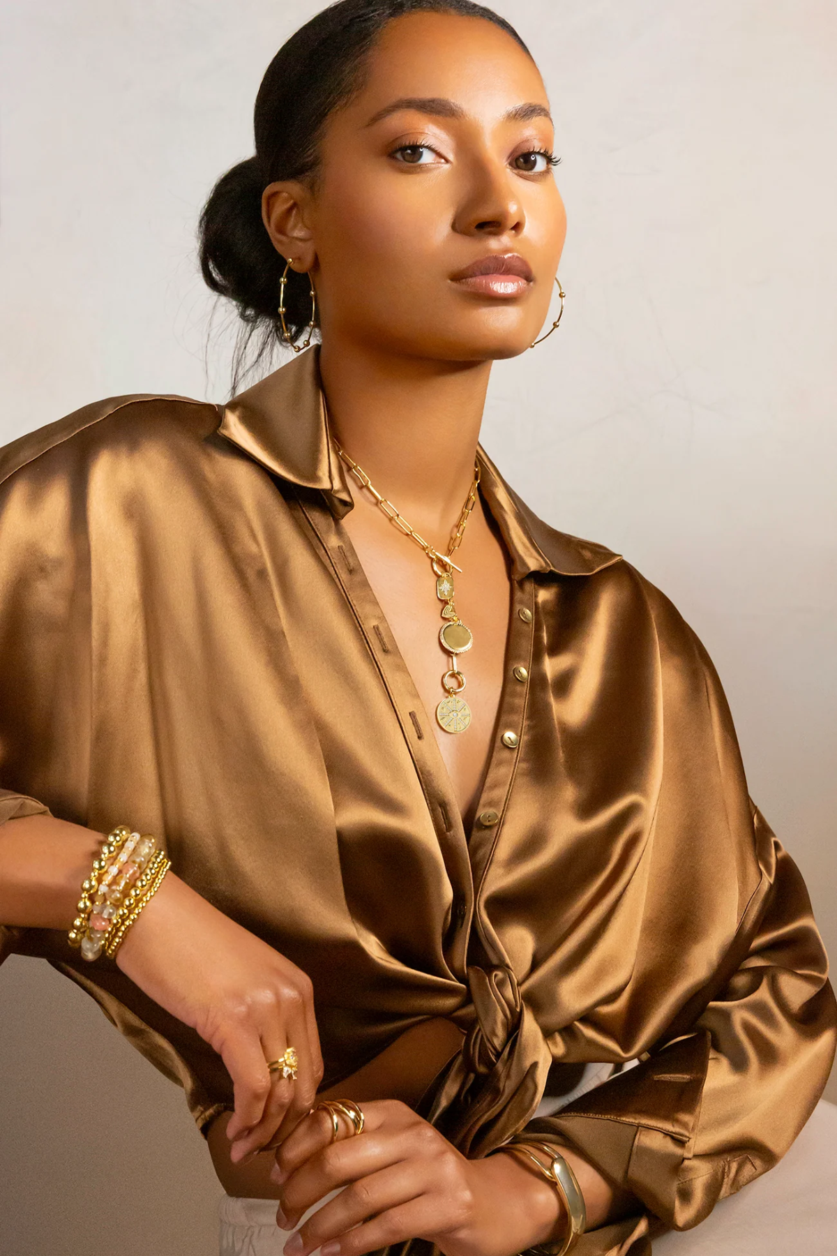 Woman wearing a gold satin shirt with jewelry against a neutral background