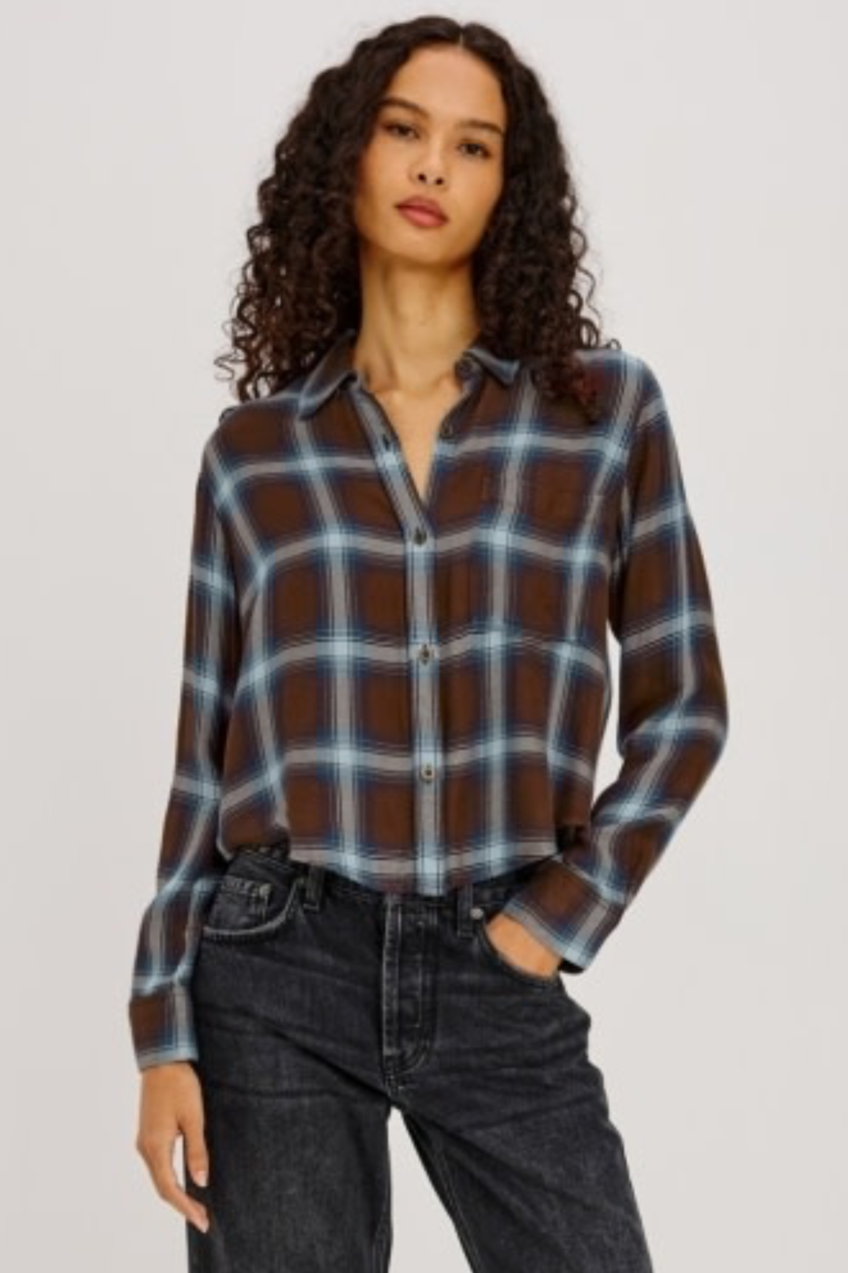 A woman modeling a brown, blue, and white plaid button-up shirt with dark wash jeans, photographed against a solid light gray background.