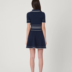 A woman viewed from the back wearing a navy blue short-sleeve mini dress with white stripe accents, paired with nude high-heeled sandals, against a light gray studio background.