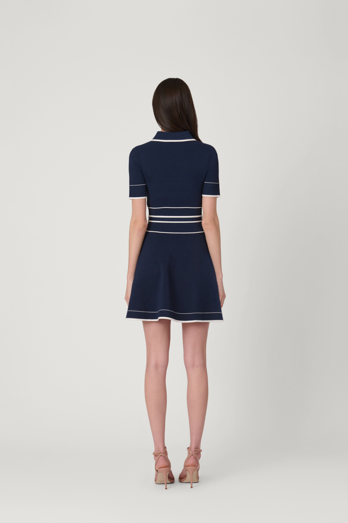 A woman viewed from the back wearing a navy blue short-sleeve mini dress with white stripe accents, paired with nude high-heeled sandals, against a light gray studio background.