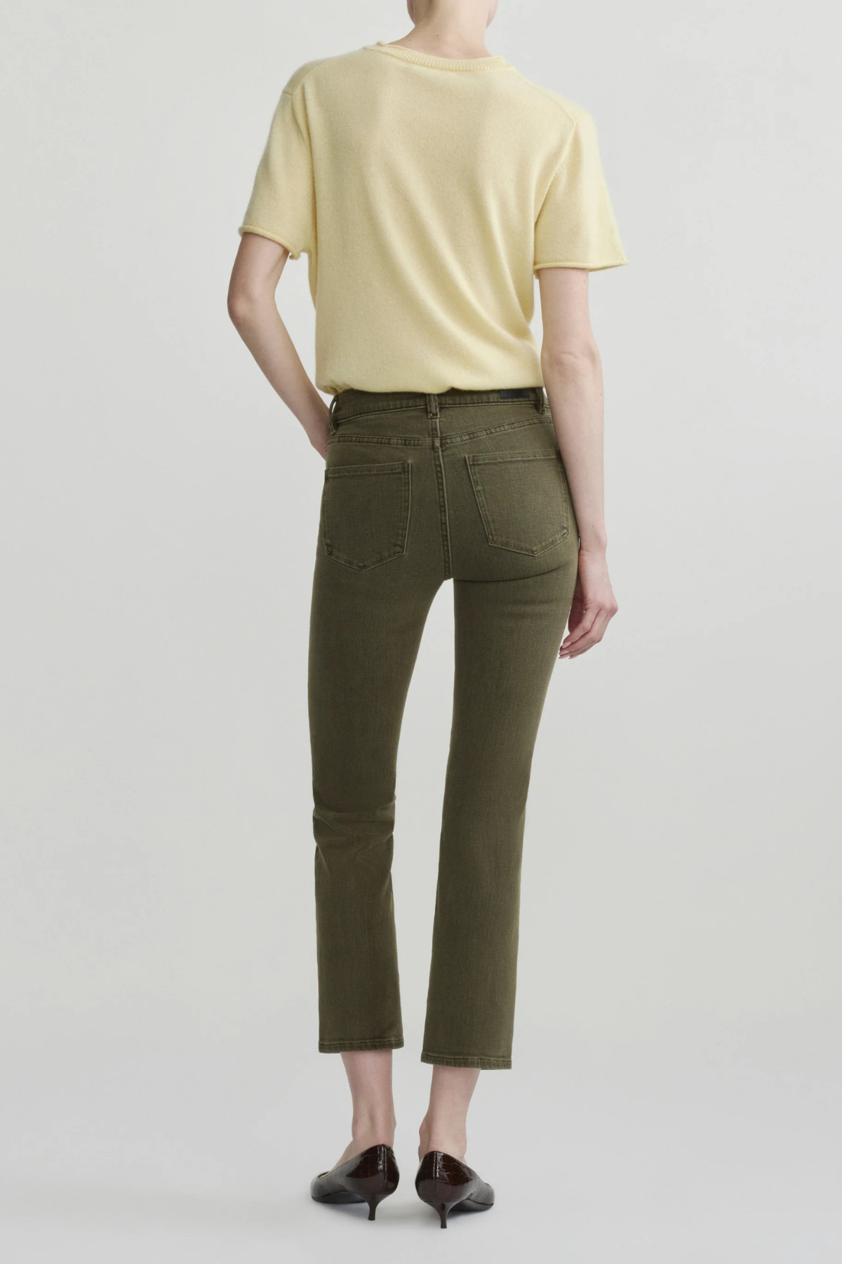 A model shown from the back wearing olive green cropped jeans, a pale yellow t-shirt, and dark brown pointed-toe flats against a light gray studio background.
