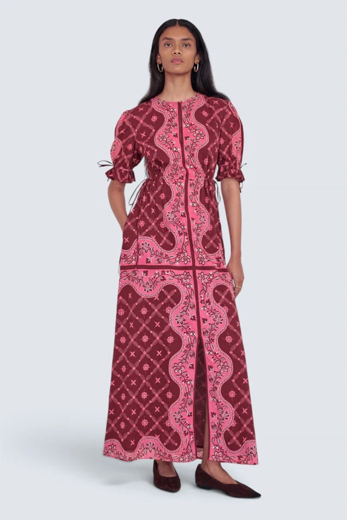 A model wearing a long-sleeved maxi dress with a pink and burgundy paisley pattern, standing against a light gray studio background.