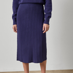 A model wearing a dark purple ribbed knit midi skirt with a matching sweater and brown loafers, standing against a neutral studio background.