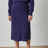 A model wearing a dark purple ribbed knit midi skirt with a matching sweater and brown loafers, standing against a neutral studio background.