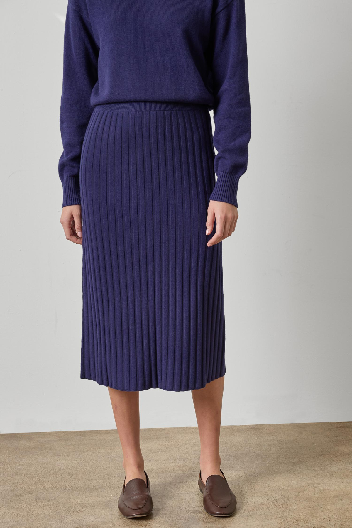 A model wearing a dark purple ribbed knit midi skirt with a matching sweater and brown loafers, standing against a neutral studio background.