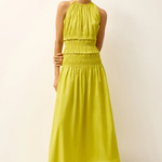 A woman modeling a sleeveless halter-neck maxi dress in a bright yellow-green color, paired with brown flat sandals, against a plain off-white studio background.