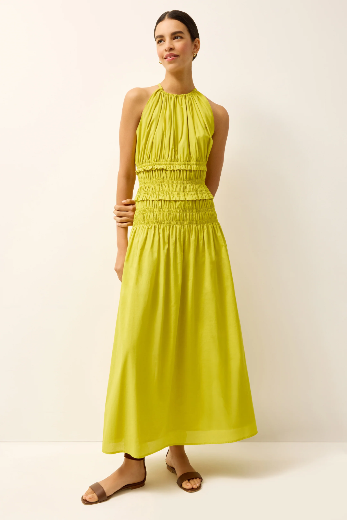A woman modeling a sleeveless halter-neck maxi dress in a bright yellow-green color, paired with brown flat sandals, against a plain off-white studio background.