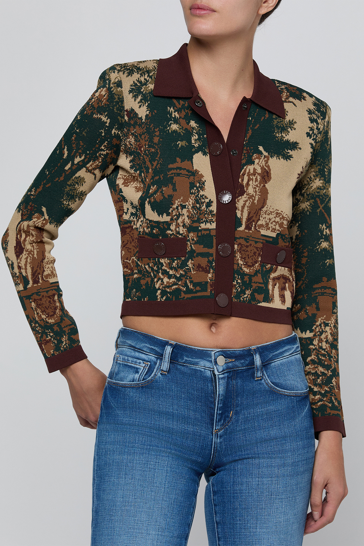 A cropped long-sleeve button-front cardigan with a dark green and beige toile-style print, worn by a model with blue jeans against a light gray studio background.