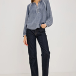 A woman modeling a light blue long-sleeve blouse with embroidered details on the chest and dark wash jeans, standing against a plain light gray studio background.