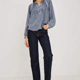 A woman modeling a light blue long-sleeve blouse with embroidered details on the chest and dark wash jeans, standing against a plain light gray studio background.