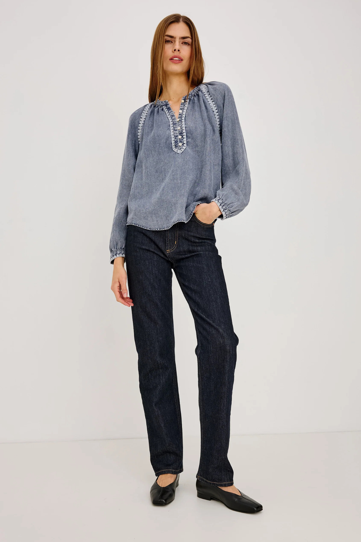 A woman modeling a light blue long-sleeve blouse with embroidered details on the chest and dark wash jeans, standing against a plain light gray studio background.