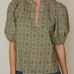A woman wearing a short-sleeve blouse with puff sleeves and a split keyhole neckline, featuring an olive green and beige geometric pattern. She is also wearing blue jeans, set against a plain beige background.