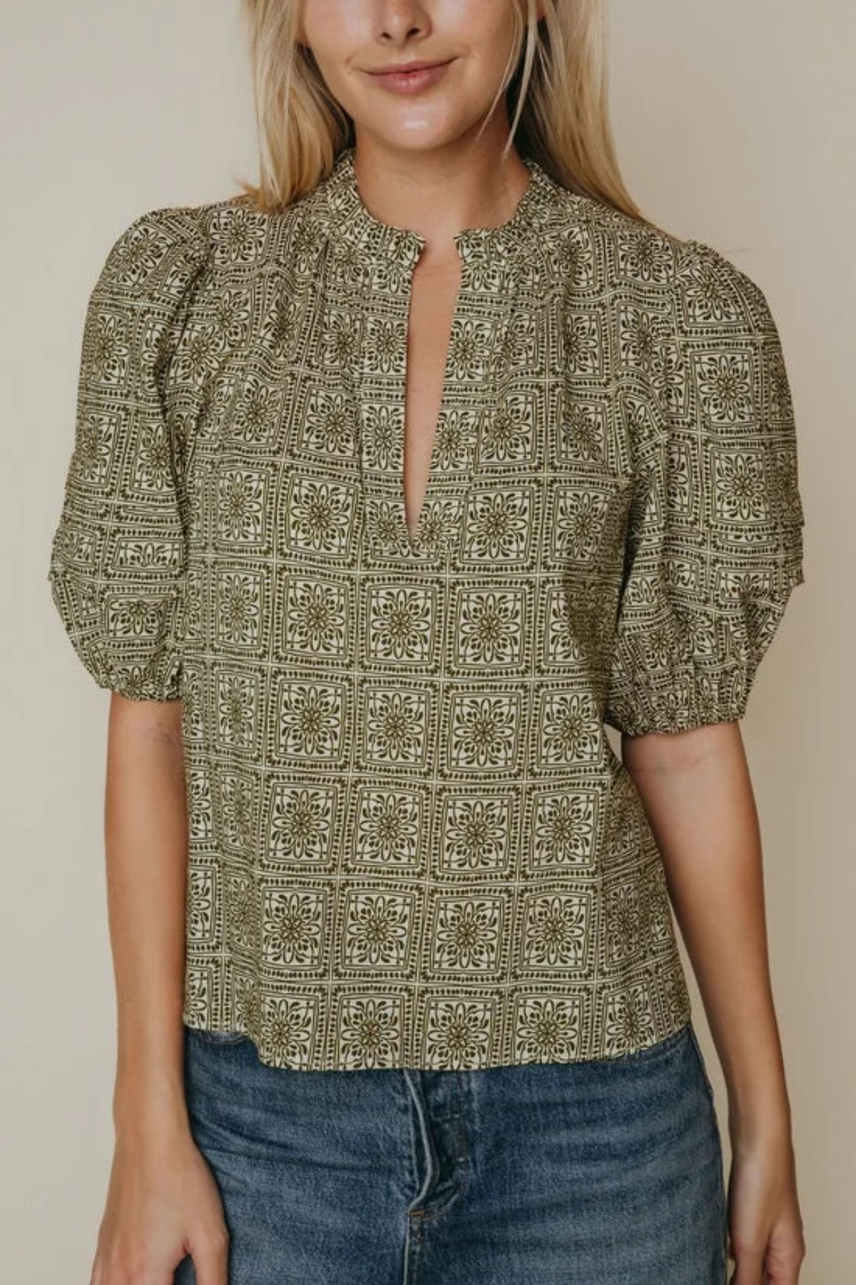 A woman wearing a short-sleeve blouse with puff sleeves and a split keyhole neckline, featuring an olive green and beige geometric pattern. She is also wearing blue jeans, set against a plain beige background.
