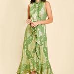 A woman modeling a sleeveless green midi dress with a gold paisley pattern, ruffled tie-neck detail, and a ruffled hem, set against a solid beige background.