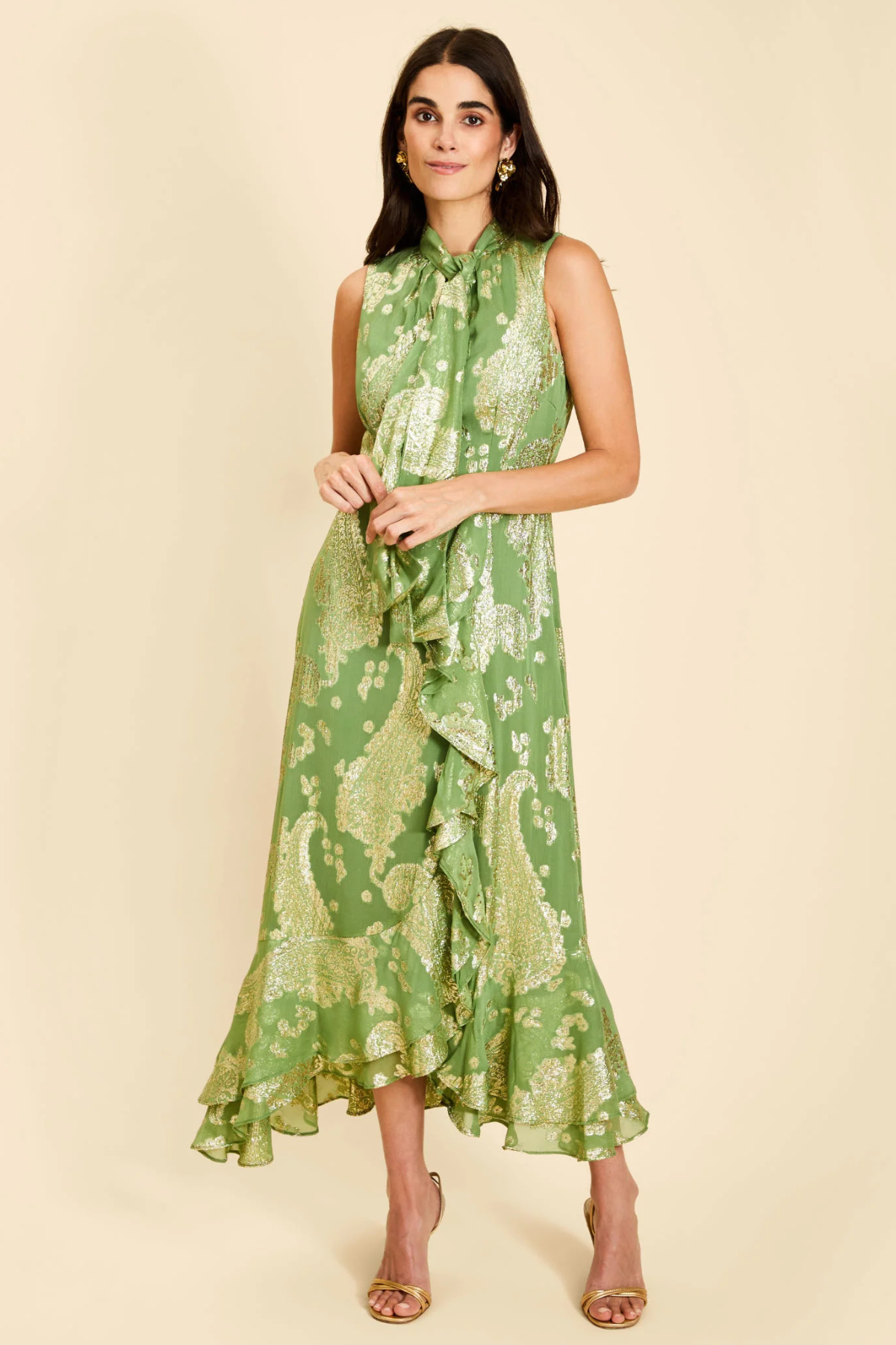 A woman modeling a sleeveless green midi dress with a gold paisley pattern, ruffled tie-neck detail, and a ruffled hem, set against a solid beige background.