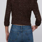 A person shown from the back wearing a dark brown textured knit sweater covered in small silver sequins, paired with blue jeans, against a light gray studio background.
