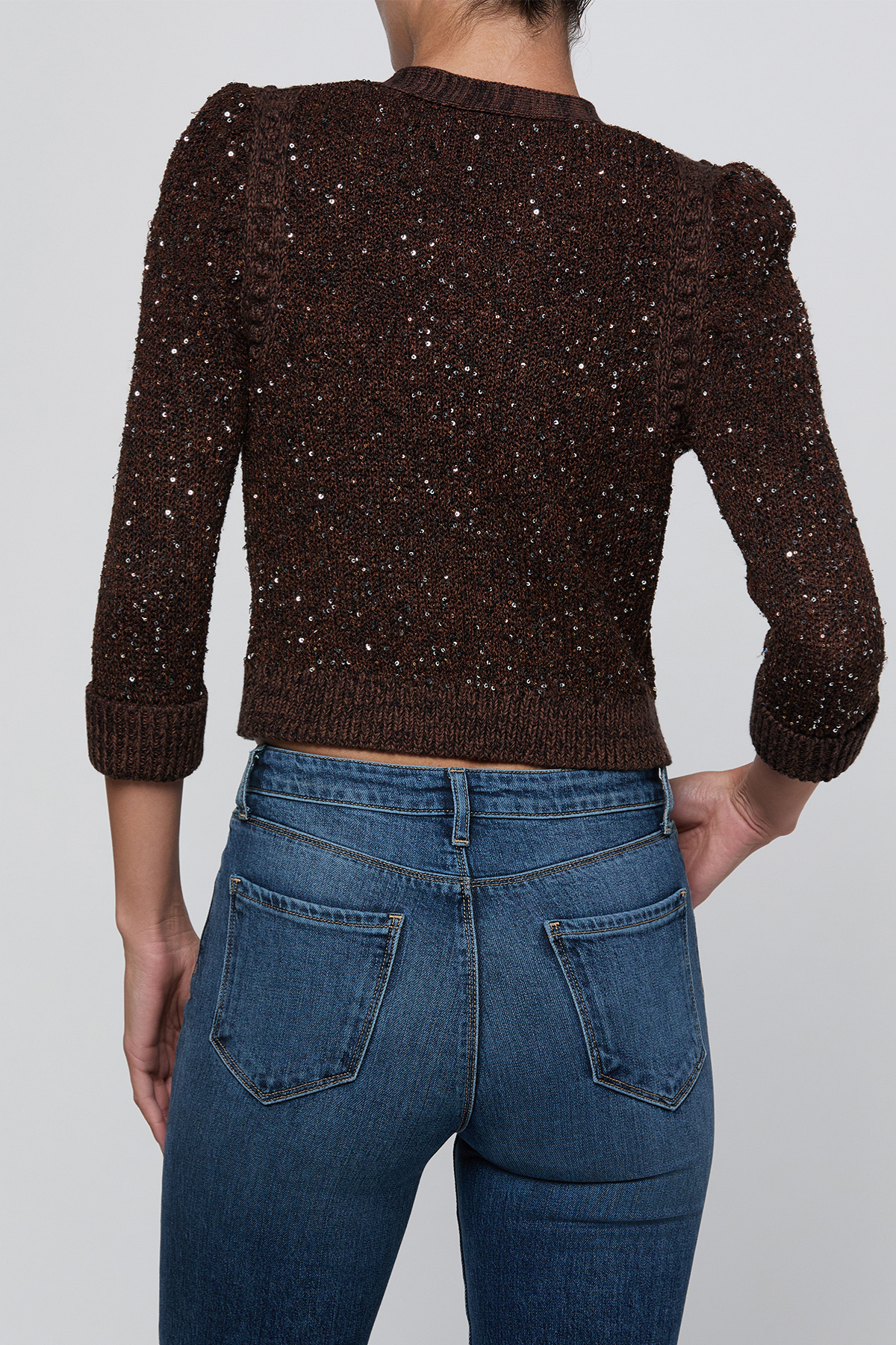 A person shown from the back wearing a dark brown textured knit sweater covered in small silver sequins, paired with blue jeans, against a light gray studio background.