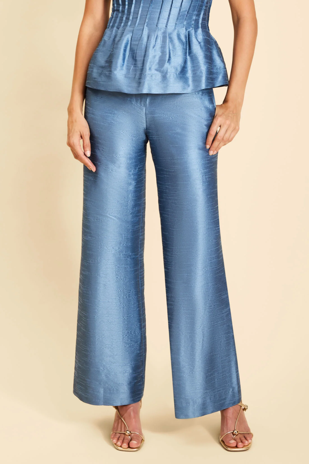 A model wearing a pair of wide-leg blue trousers with a subtle sheen, paired with a matching blue peplum top and metallic sandals, set against a solid beige background.