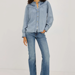 A model stands against a plain light gray studio background wearing a light blue denim button-up shirt with embroidered detailing on the collar, matching light blue jeans, and dark brown pointed-toe heeled shoes.