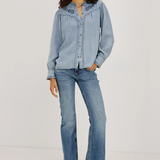A model stands against a plain light gray studio background wearing a light blue denim button-up shirt with embroidered detailing on the collar, matching light blue jeans, and dark brown pointed-toe heeled shoes.