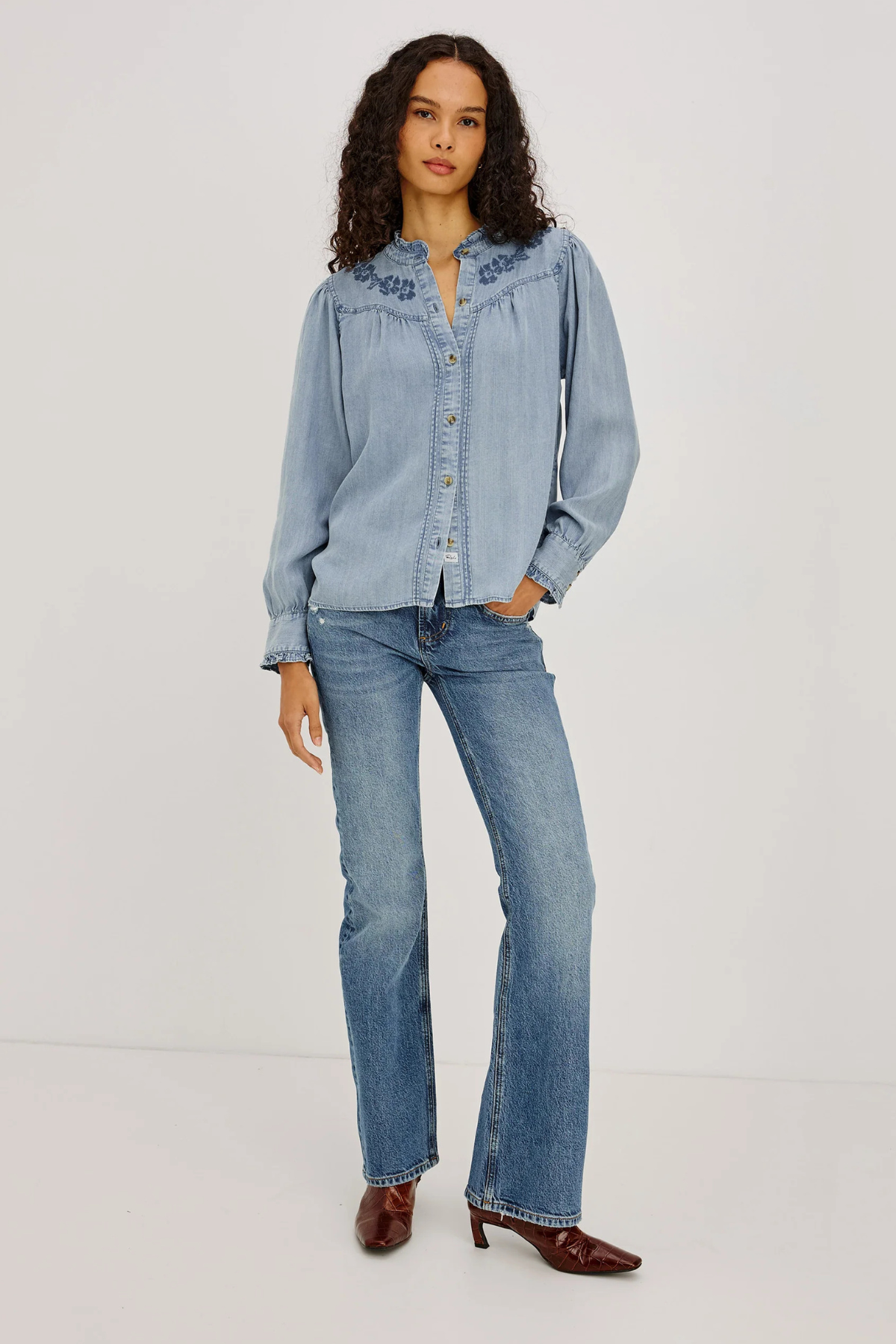 A model stands against a plain light gray studio background wearing a light blue denim button-up shirt with embroidered detailing on the collar, matching light blue jeans, and dark brown pointed-toe heeled shoes.