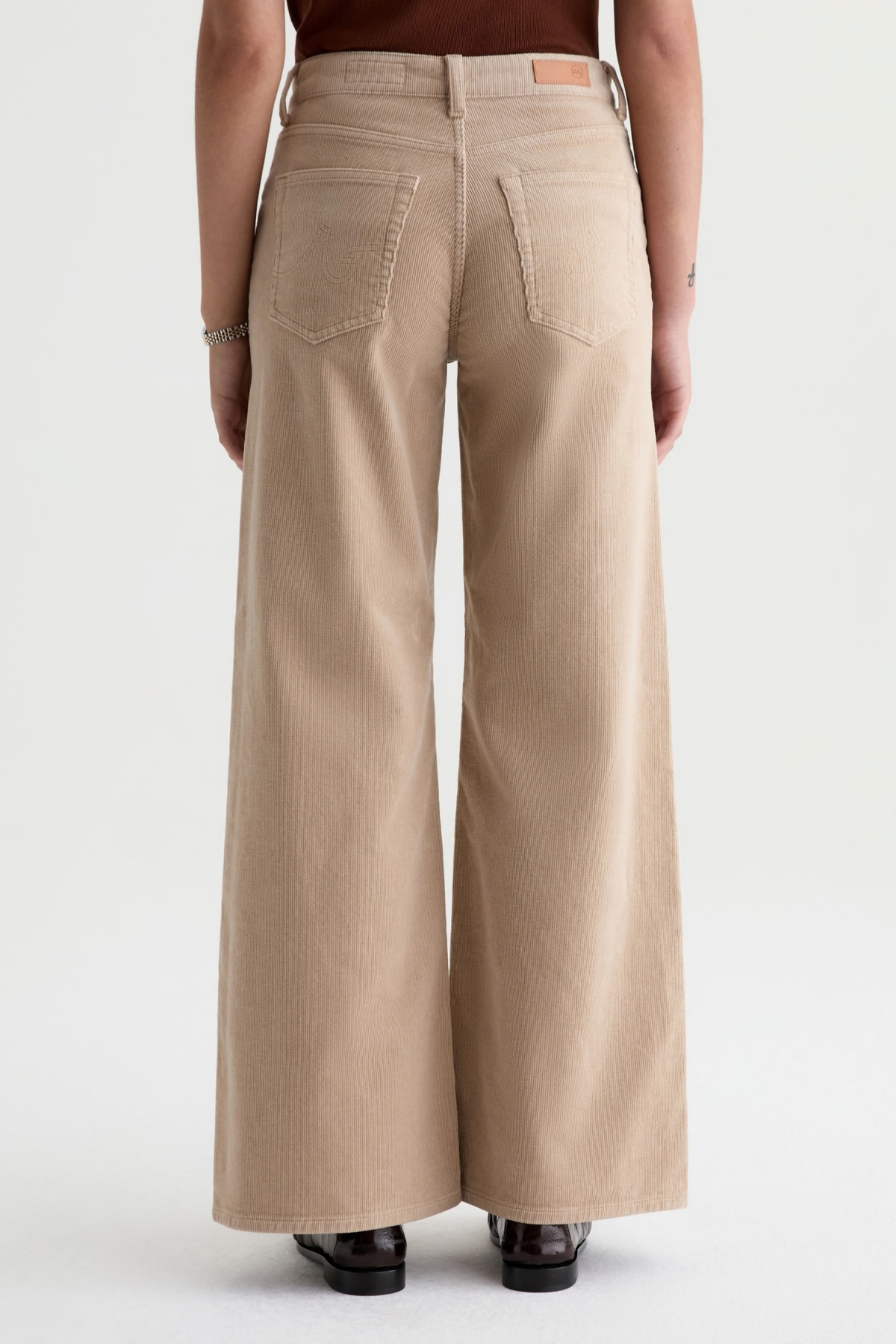 A person shown from the back wearing light beige wide-leg jeans with a brown top and black shoes, photographed against a plain light gray studio background.