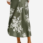 A woman wearing a midi-length olive green satin skirt with a white floral print, styled with a cream long-sleeve top and black strappy high-heeled sandals against a light gray background.
